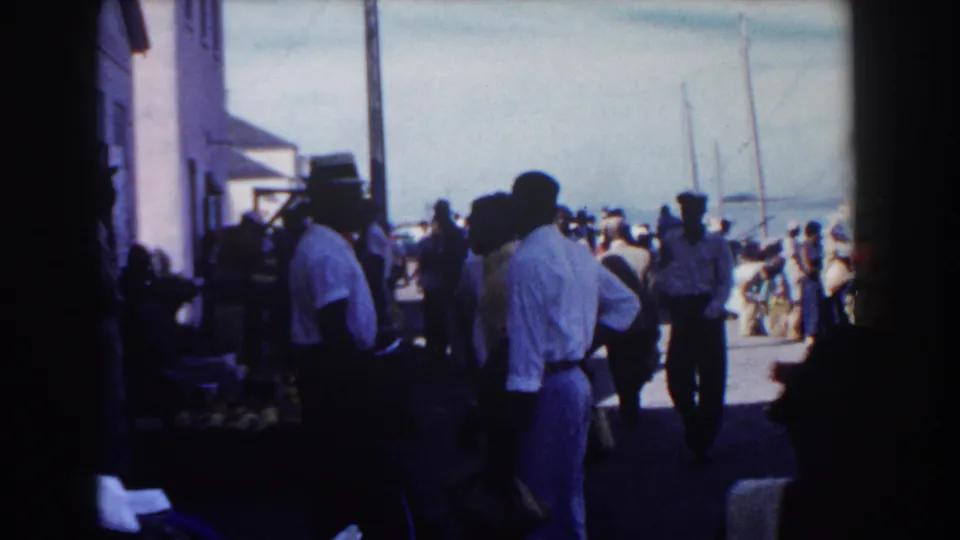 Handheld 8mm view of a busy Nassau pier, with foreground figures in shadow and harbor activity beyond.