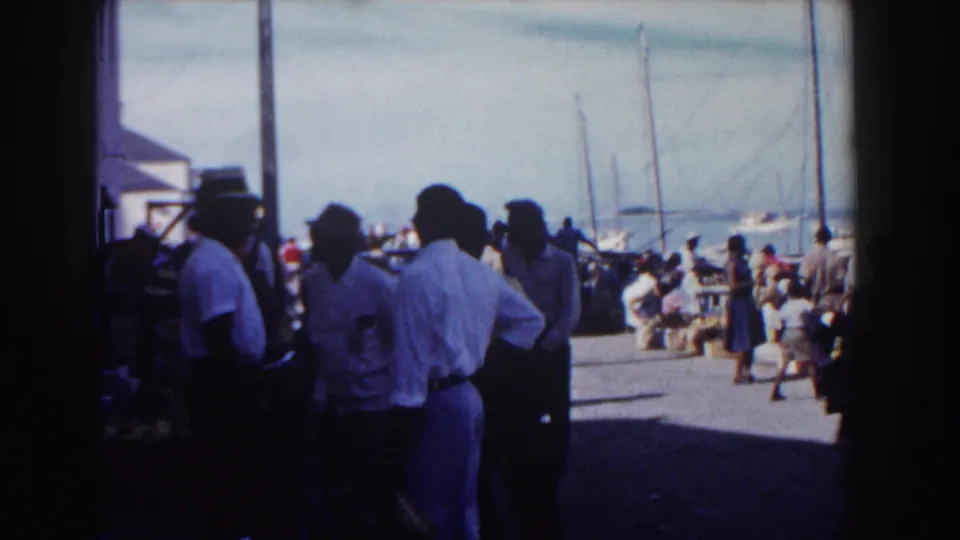 1961 Nassau pier view with people gathered in partial shade, open dock space, and sailboats in the background.