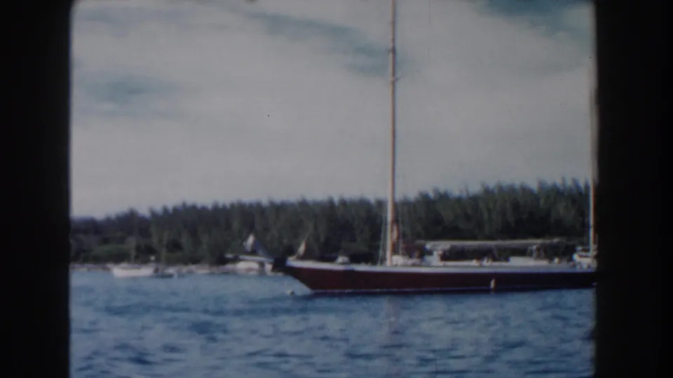 1961 Nassau frame with a dark-hulled sailboat gliding past blue water and a forested shore.
