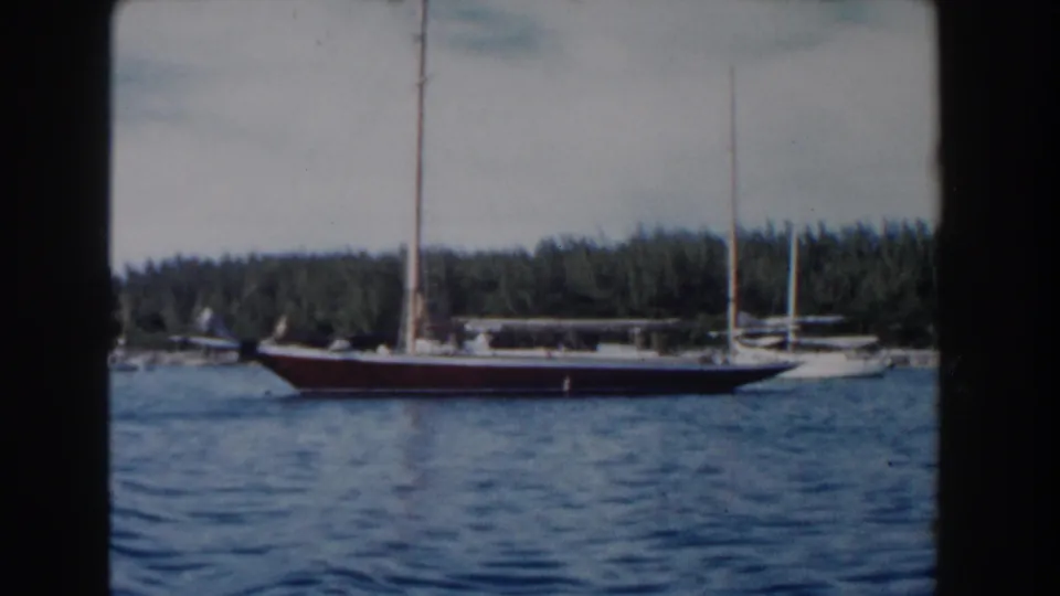 1961 Nassau home-movie still of a sailboat crossing in front of a tree-lined coastal background.