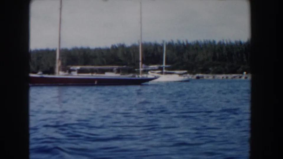 1961 Nassau frame showing a sailboat moving along a wooded coast, with open water filling the foreground.