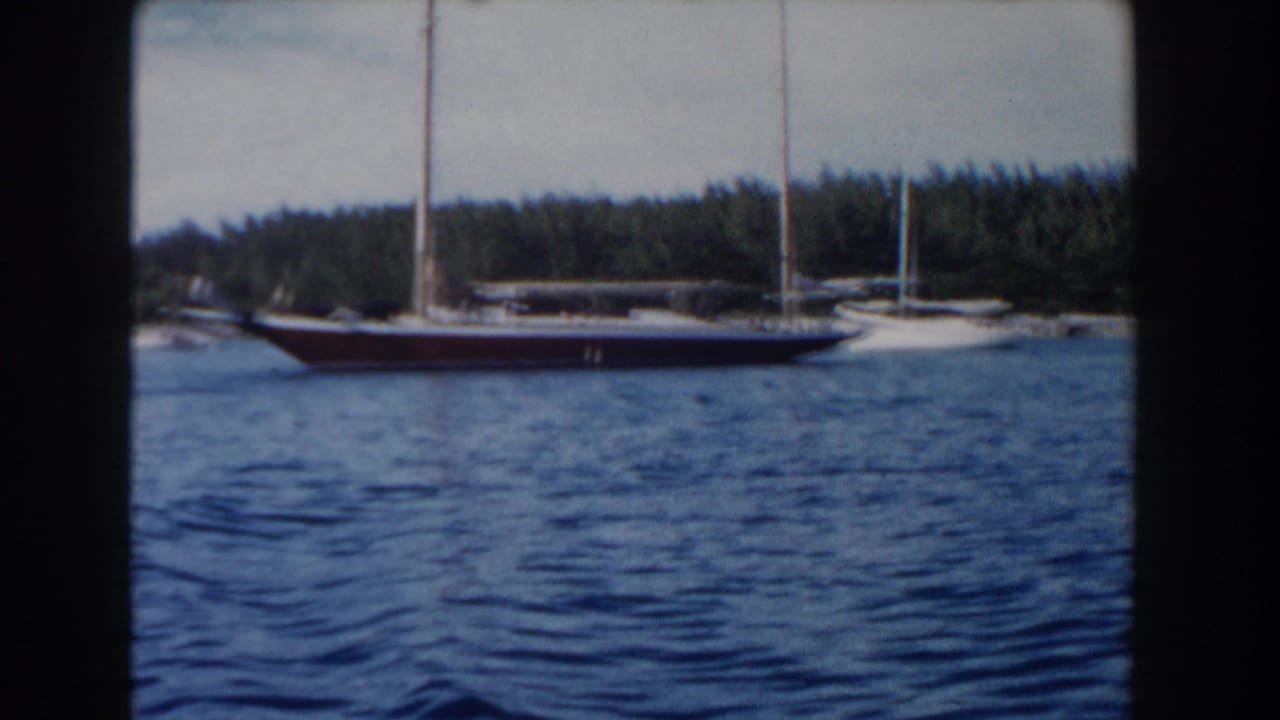 1961 Nassau home-movie frame showing a red-hulled sailboat crossing blue harbor water near a tree-lined shore.