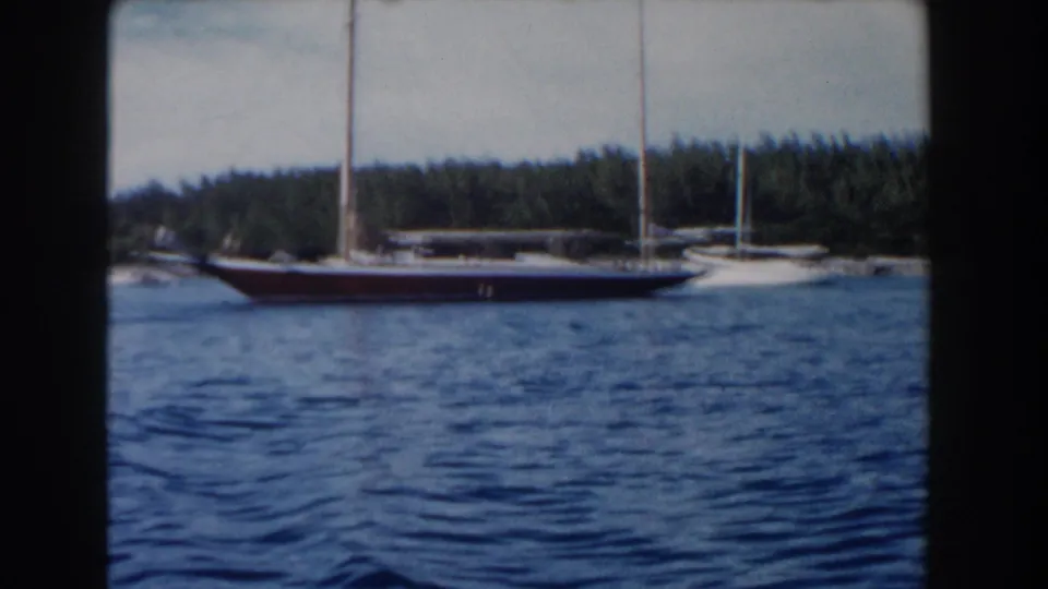 1961 Nassau home-movie frame showing a red-hulled sailboat crossing blue harbor water near a tree-lined shore.
