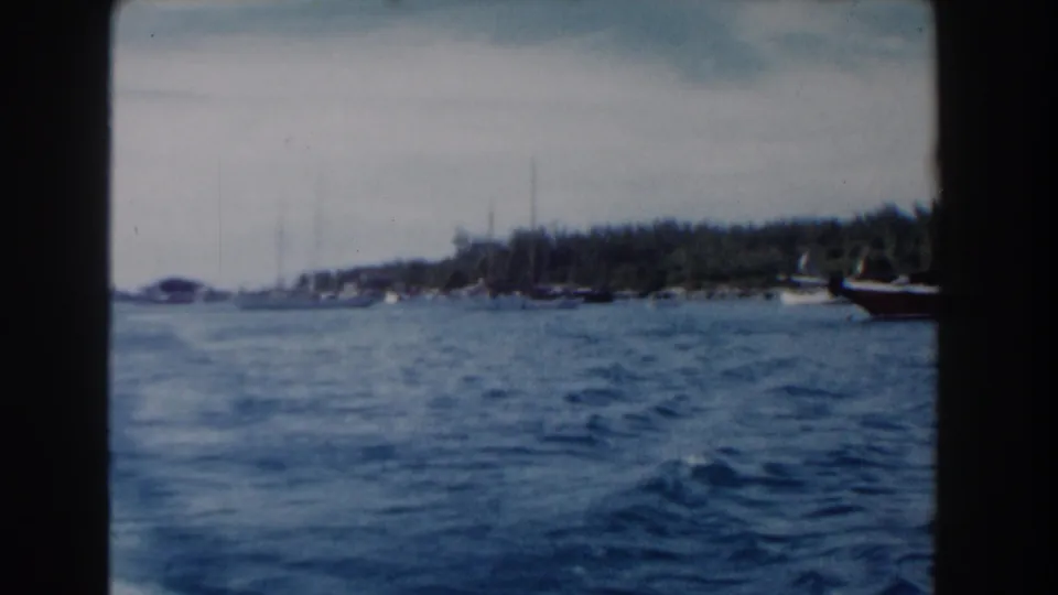 1961 Nassau home-movie frame showing a wider harbor view with small boats, blue water, and a low green shoreline.