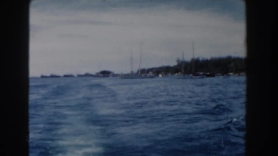 1961 Nassau harbor frame showing open water, distant sailboats, and a tree-lined shore under a pale sky.