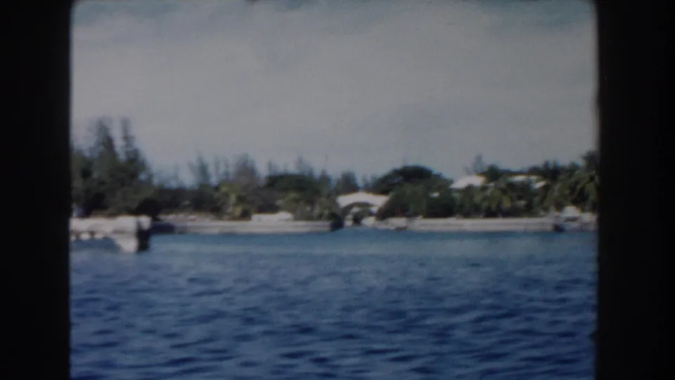 Nassau harbor shoreline in 1961 with blue water, palms, low buildings, and a small bridge or arched structure in the distance.