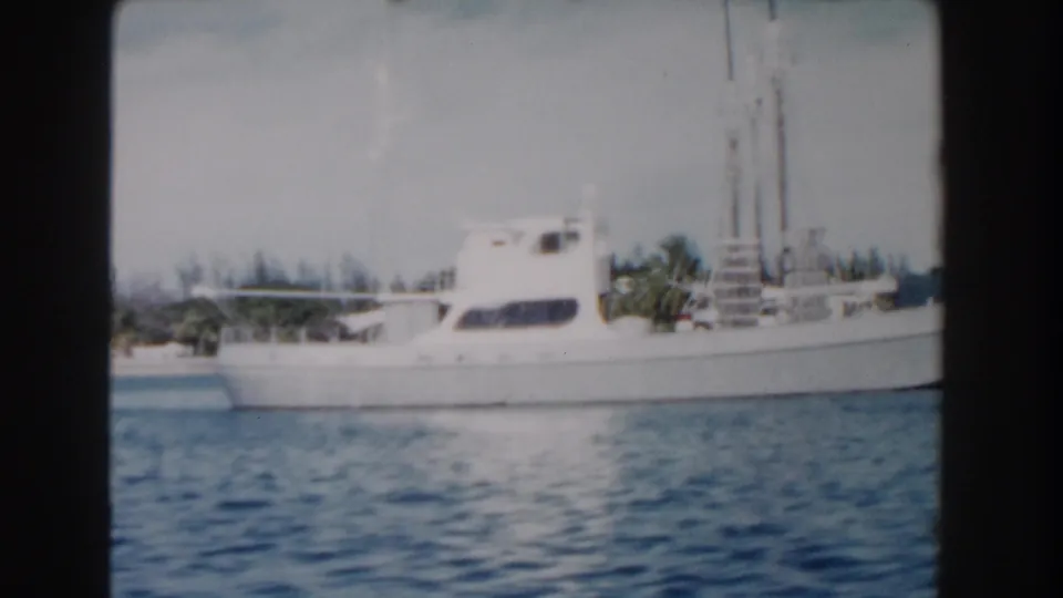 1961 Nassau harbor frame showing a white yacht on blue water with palms and low shoreline structures in the background.
