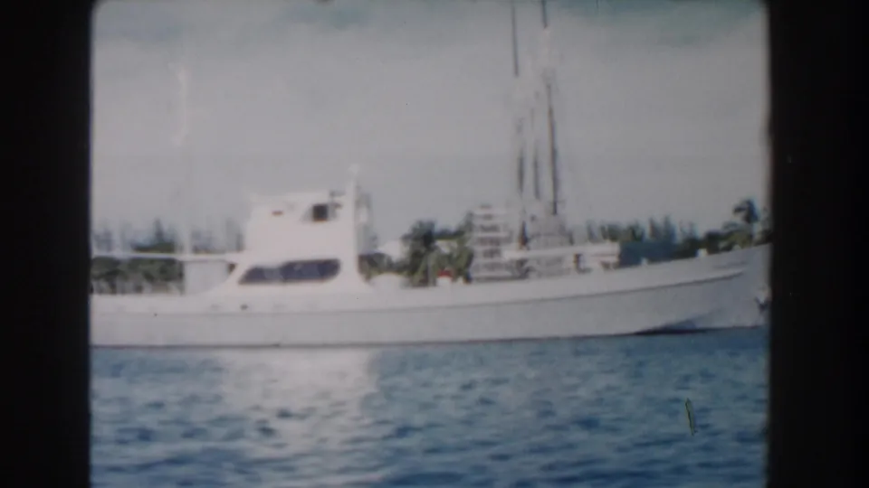 White cabin boat seen from the side on blue Nassau water in 1961 home-movie footage, with palms and shore in the background.