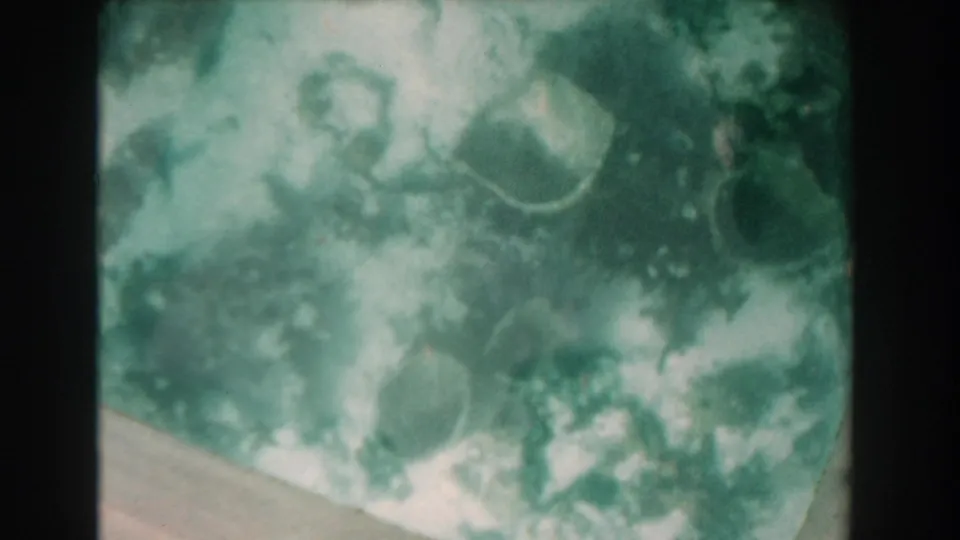 1961 Nassau underwater footage frame with diffuse light, green water, and scattered coral or rocks near a pale seafloor slope.
