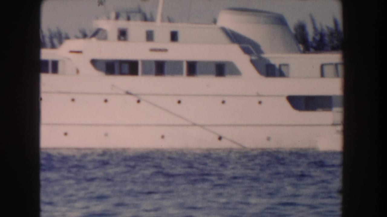 1961 Nassau home-movie frame showing the side of a large white yacht across calm blue water, with shoreline trees in the background.
