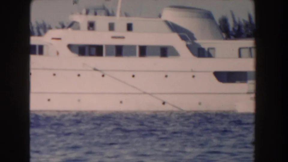 1961 Nassau home-movie frame showing the side of a large white yacht across calm blue water, with shoreline trees in the background.