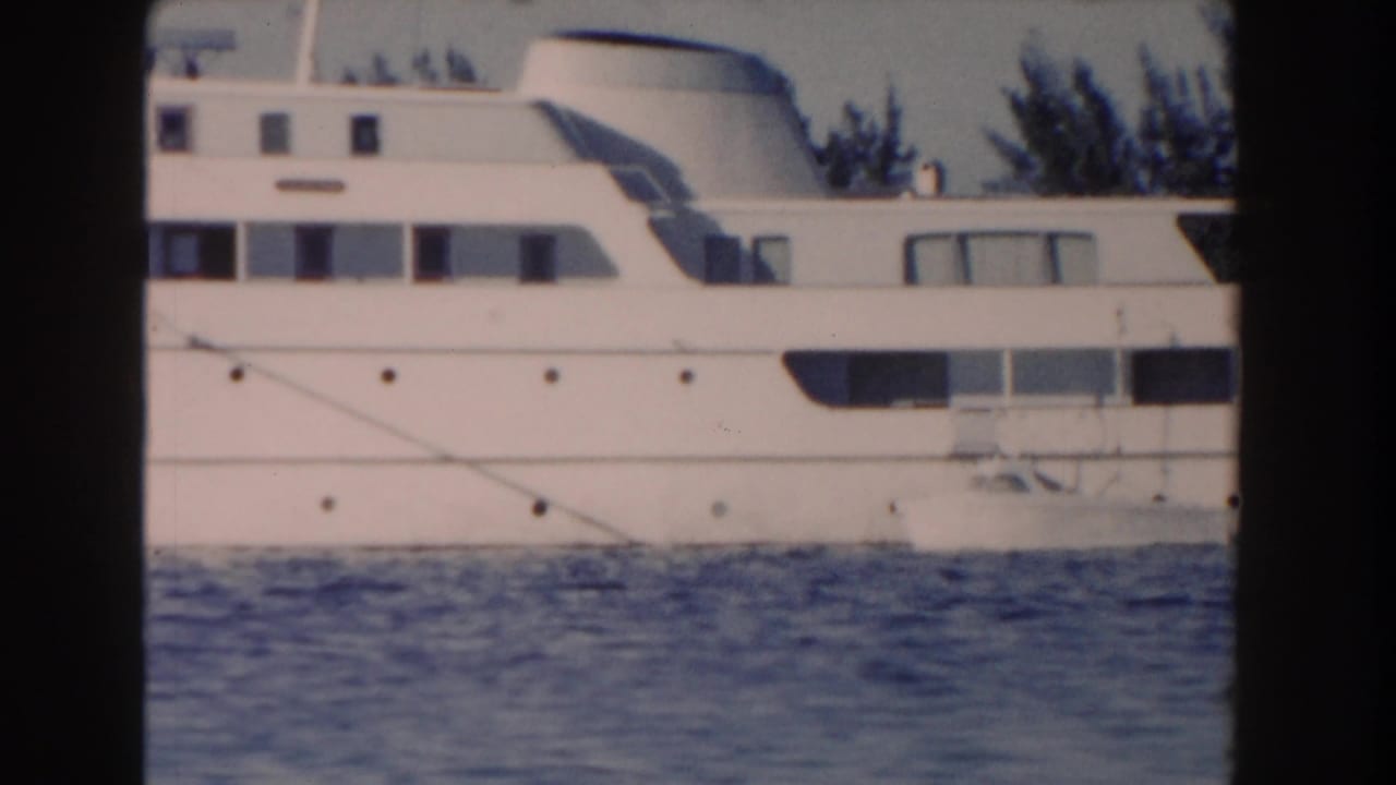 Side view of a white yacht on blue water with a tropical shoreline behind it.