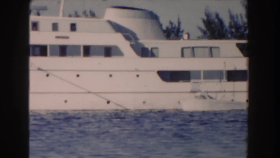 1961 Nassau frame showing the yacht broadside on calm water, with cabin windows, railings, and trees along the shore.