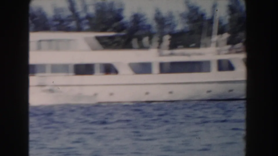 Soft 8mm frame from Nassau showing the yacht's side and cabin windows across blue water, with tropical trees behind it.