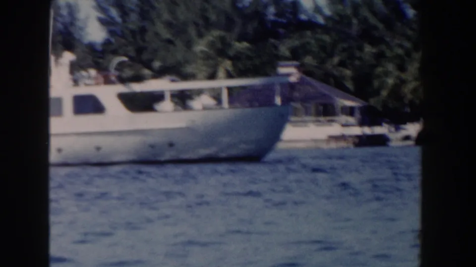 1961 Nassau frame showing the forward section of the white yacht, including the bow, windows, railings, water, and shoreline trees.