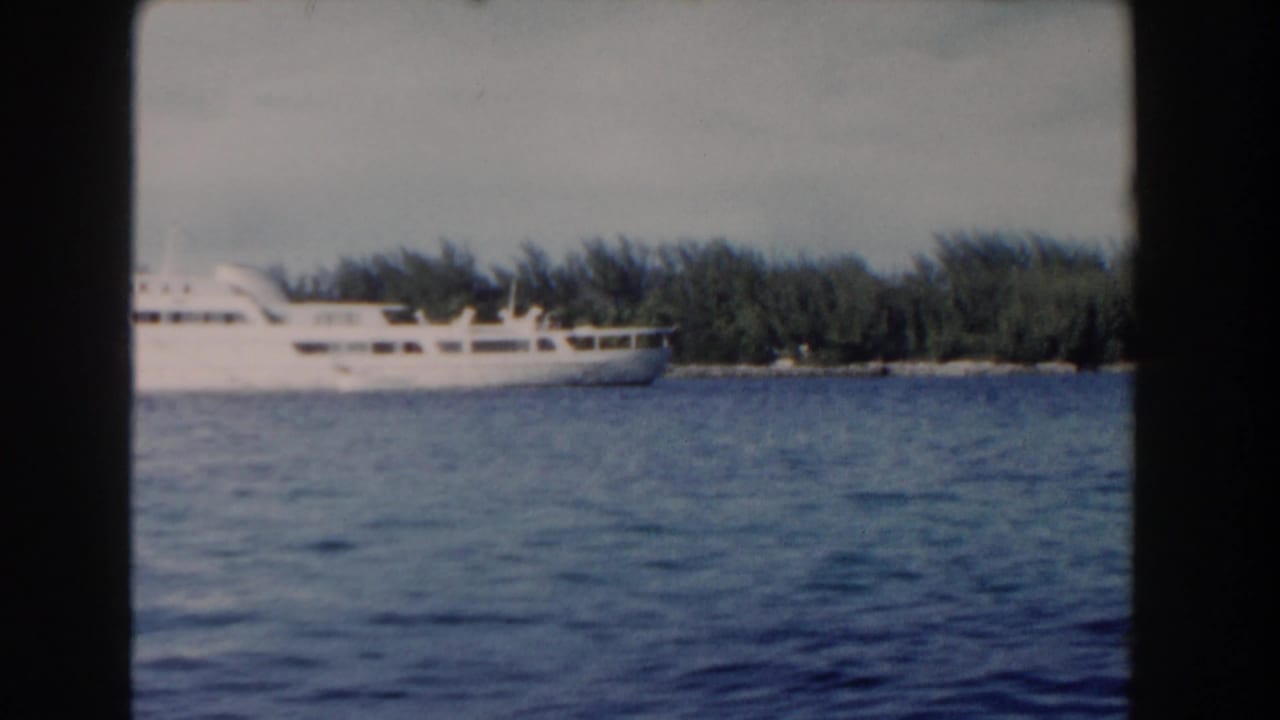 White cruise ship seen across Nassau harbor in 1961 home-movie footage, with blue water and a low tropical shoreline behind it.
