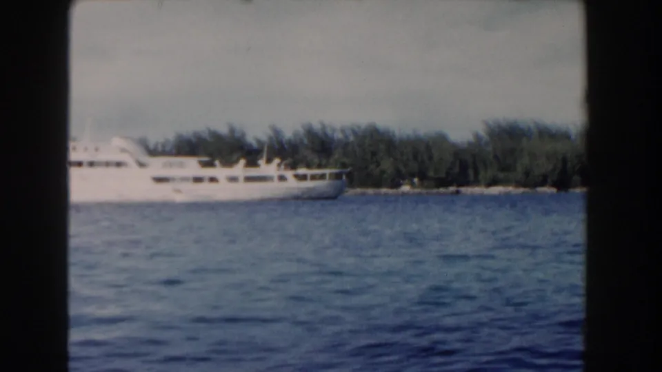White cruise ship seen across Nassau harbor in 1961 home-movie footage, with blue water and a low tropical shoreline behind it.