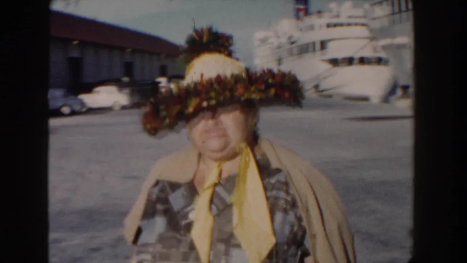 1961 Nassau dockside frame showing an older woman in a large floral hat, with the cruise ship and parked cars behind her.