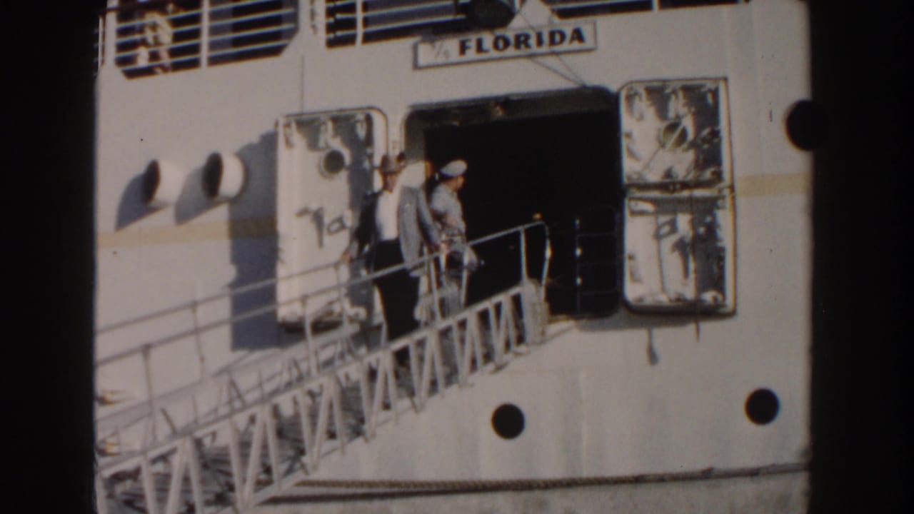 Man in a light suit and hat waves from a ship gangway beside an open doorway.
