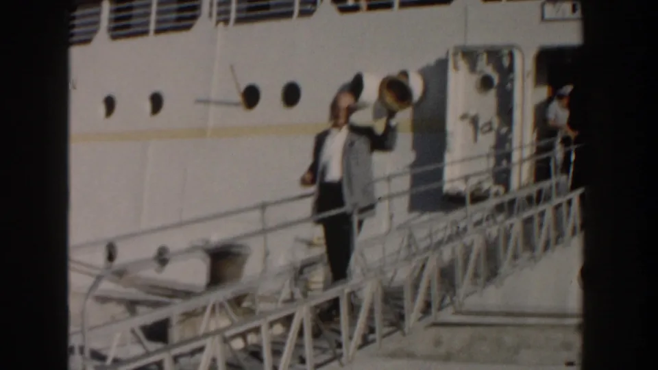 The man lifts his fedora in greeting on the ship gangway, with portholes and the open passenger doorway visible behind him.