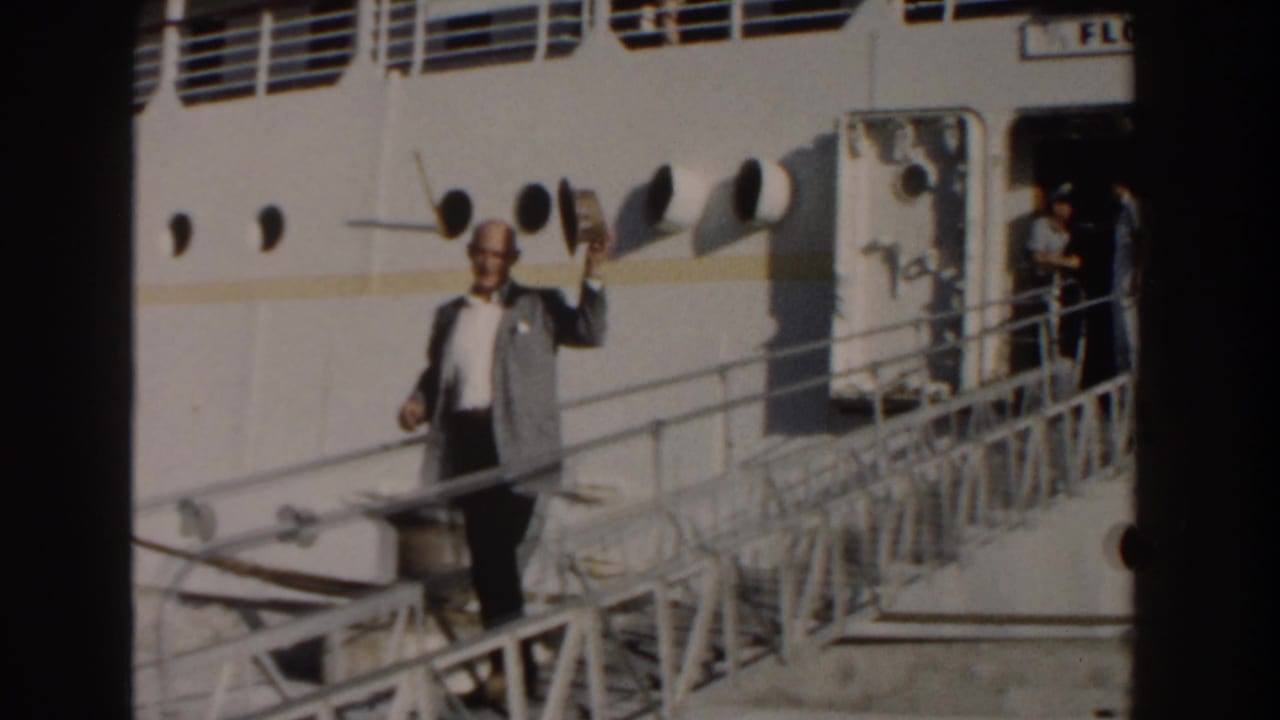 Man in a grey jacket waves a straw hat while stepping down a ship gangway.
