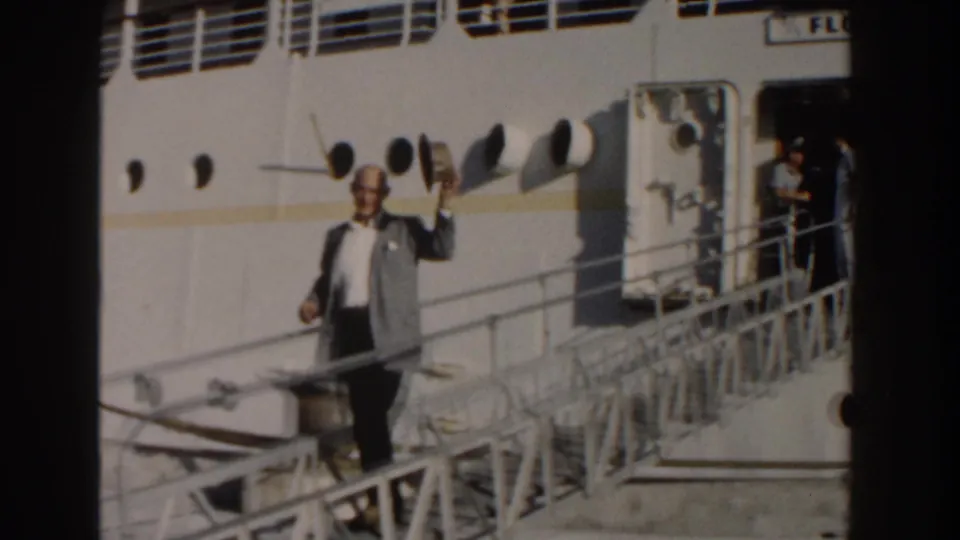 In Nassau, Bahamas, 1961, a man in a grey jacket waves a straw hat from a white gangway beside a ship hull.