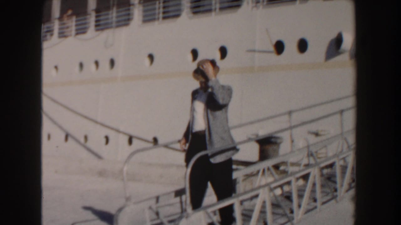 Man on the gangway holds his hat near his head beside the ship hull.