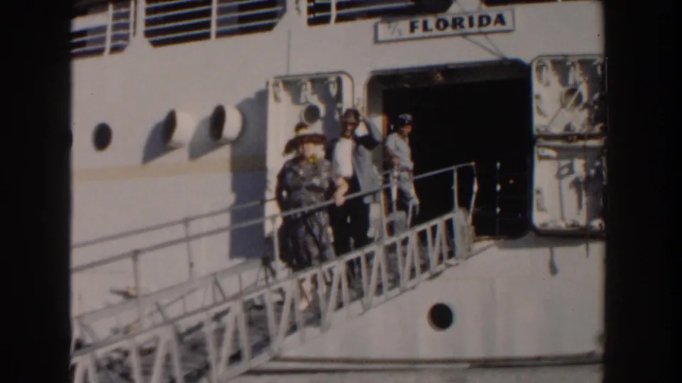 In this 1961 Nassau frame, the man lifts or waves his hat while standing with a woman on the ship gangway.