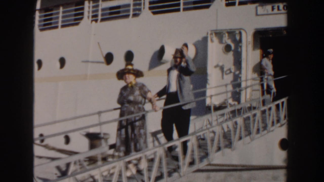 Woman in a wide-brimmed hat waves while descending a ship gangway with a man behind her.