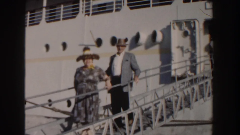 The couple continues down the gangway in front of the cruise ship hull, with portholes and deck railings visible in the 8mm frame.