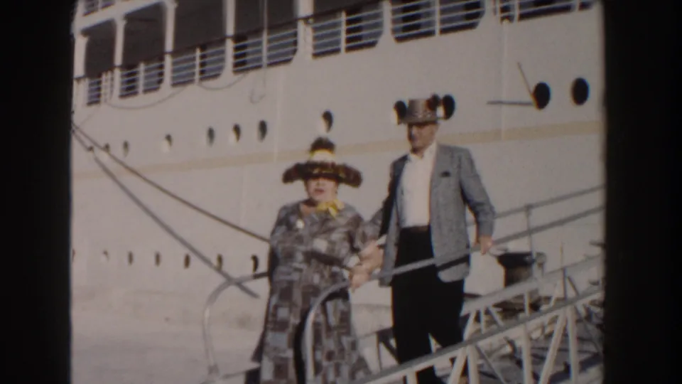 In Nassau, Bahamas, an elderly couple in 1960s travel clothing descends a gangway from a large white cruise ship.