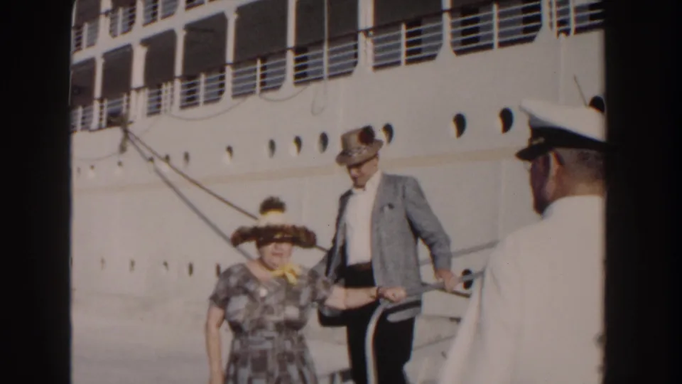 The elderly couple continues down the cruise ship gangway as a white-uniformed crew member stands near the dockside rail.