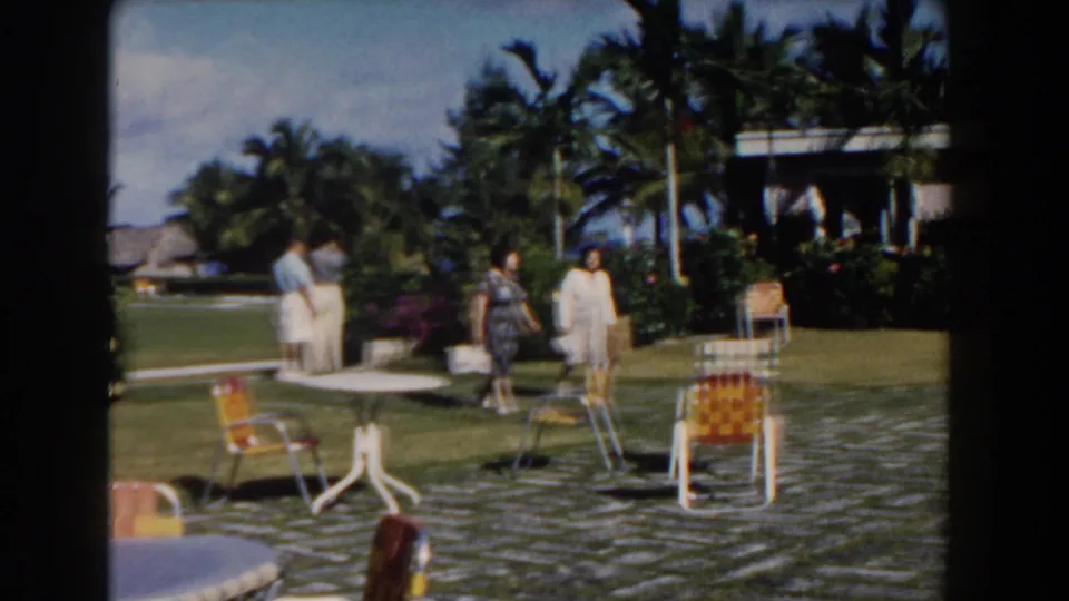 At a Nassau resort setting, guests move across a manicured lawn with palm trees, patio chairs, tables, and a shaded building nearby.
