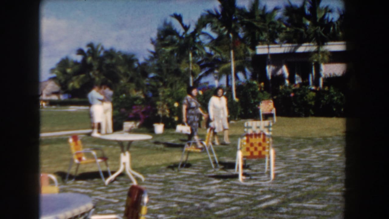 Nassau, Bahamas 1961 home-movie frame showing adults gathered in a bright courtyard with palms, white tables, and woven chairs.