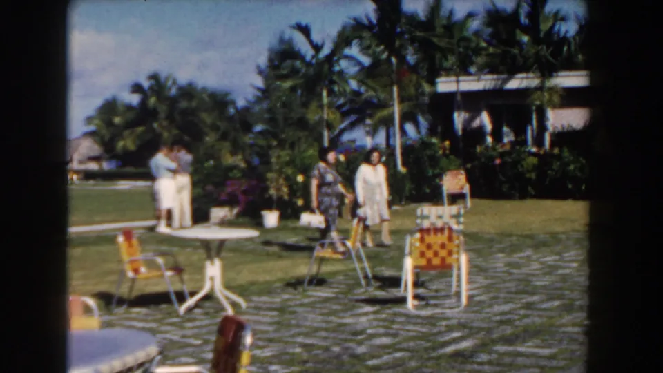 Nassau, Bahamas 1961 home-movie frame showing adults gathered in a bright courtyard with palms, white tables, and woven chairs.