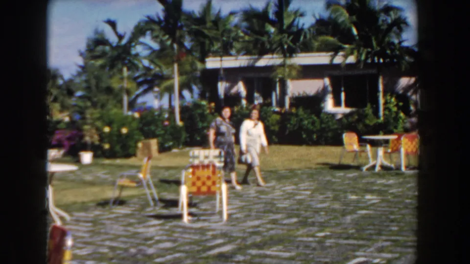 Two women in period dresses walk across a grey stone courtyard in Nassau, Bahamas, with tropical plants and patio chairs around them.