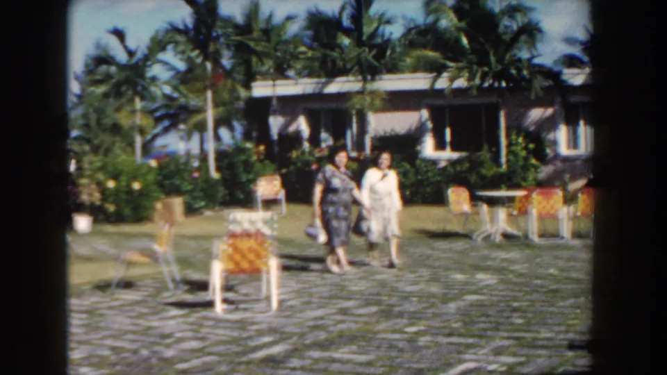 Two women walk through a sunny 1961 Nassau courtyard, framed by woven chairs, white tables, palms, and a low modern house.