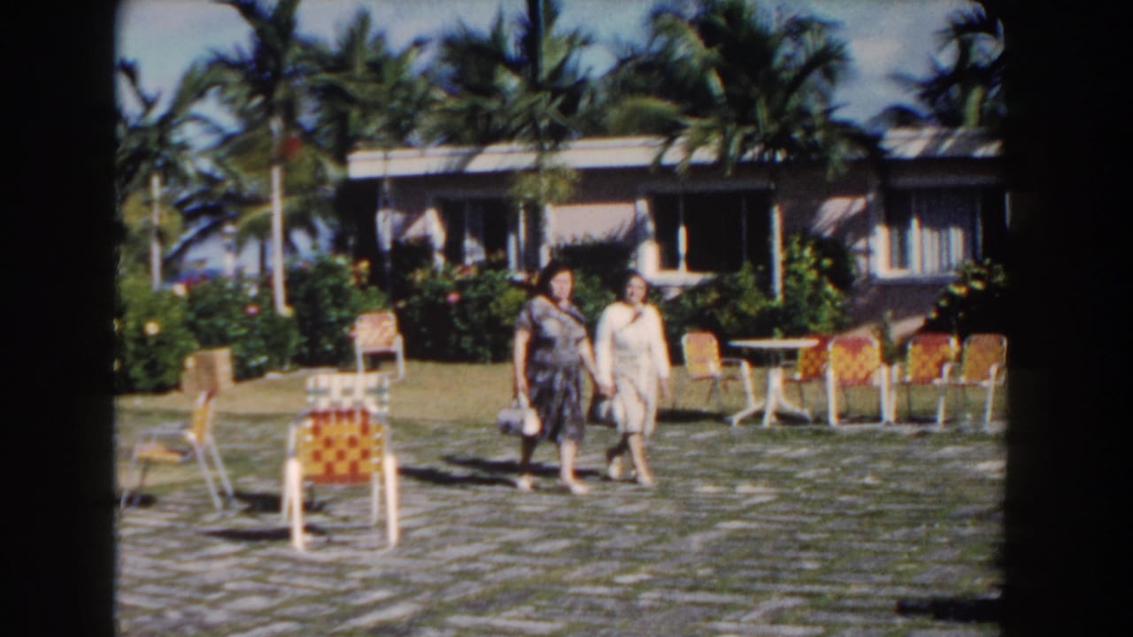 Two women walk across a paved courtyard in front of palm trees and a low resort building.