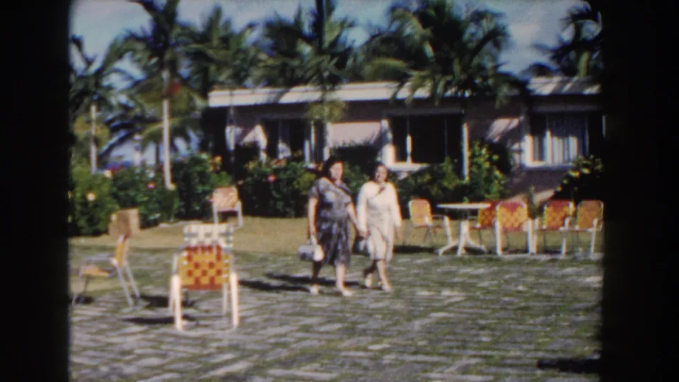 Two women in classic dresses walk together through a Nassau resort courtyard in 1961, with palm trees and patio chairs behind them.