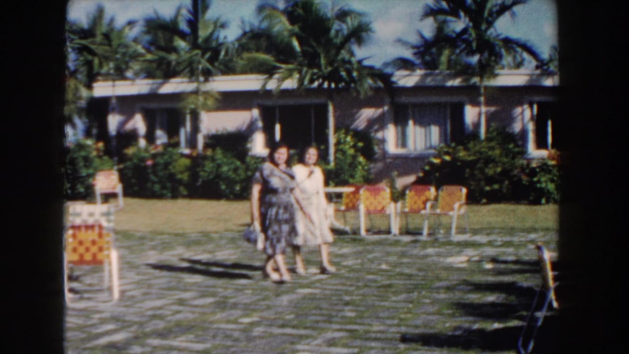 Two women stand or walk together near outdoor chairs, with palms and a resort building behind them.