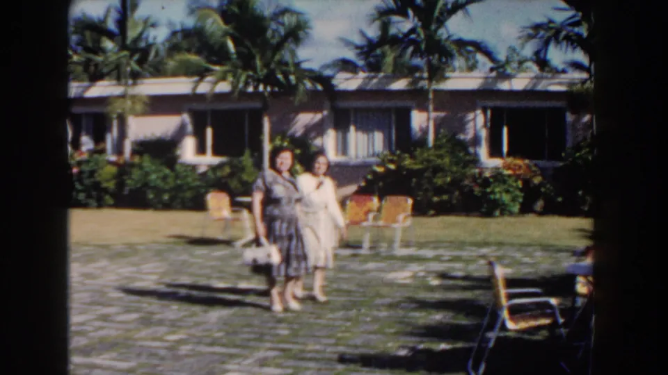 Two women in 1960s dresses approach the camera in a Nassau resort courtyard, with palm trees, windows, lawn, and outdoor seating visible.