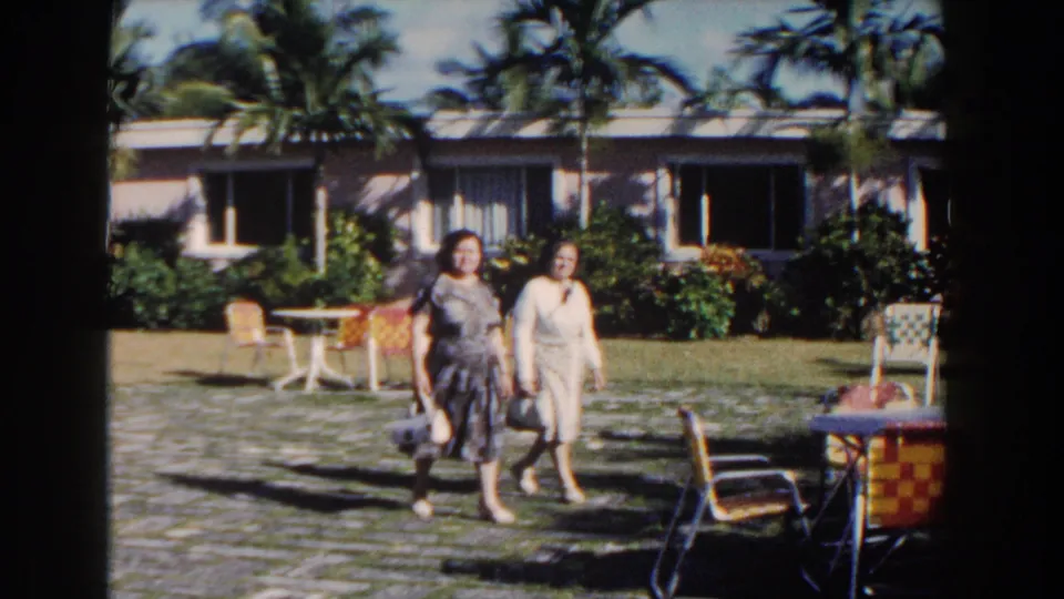 Two women walk past the camera in a 1961 Nassau resort courtyard, with colorful woven seating, palms, shrubs, and a low building behind them.
