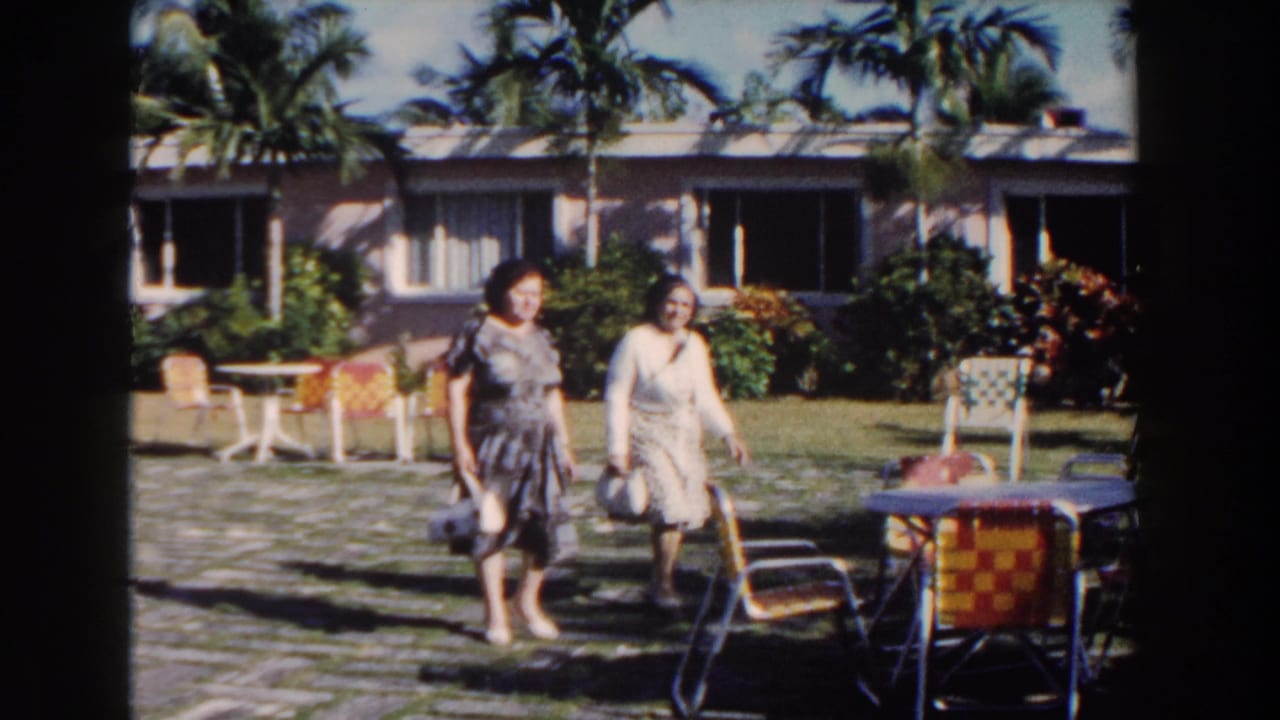 Two women walk through a sunny resort courtyard past patio chairs and tables.