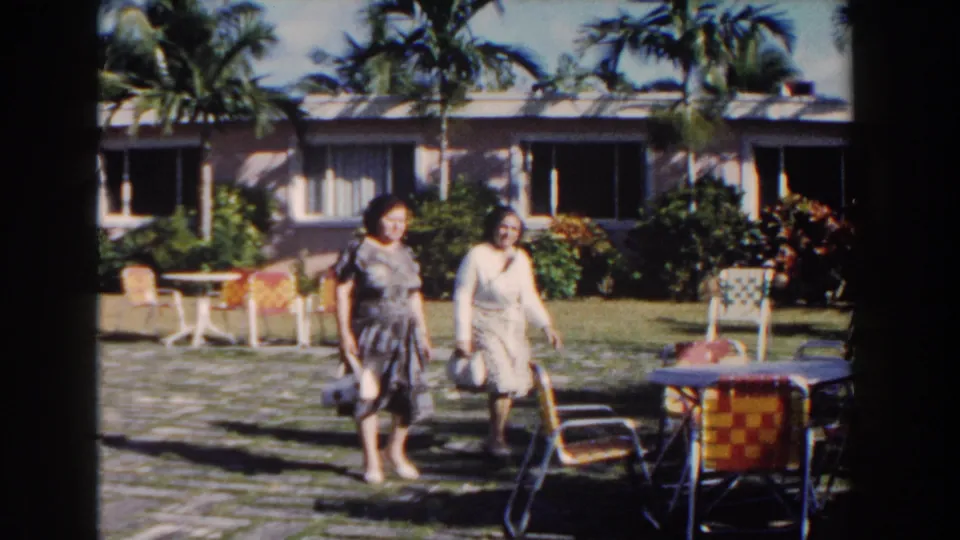 In Nassau, Bahamas in 1961, two women walk together through a tropical resort courtyard with woven chairs, tables, palms, and a low building behind them.