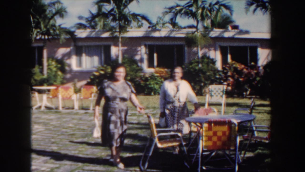 Two women approach the camera near colorful patio chairs in a tropical courtyard.