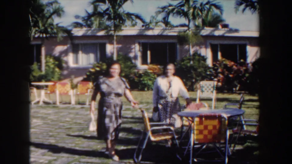 The two women continue toward the camera in the 1961 Nassau resort courtyard, carrying small bags as palm trees and a low building fill the background.