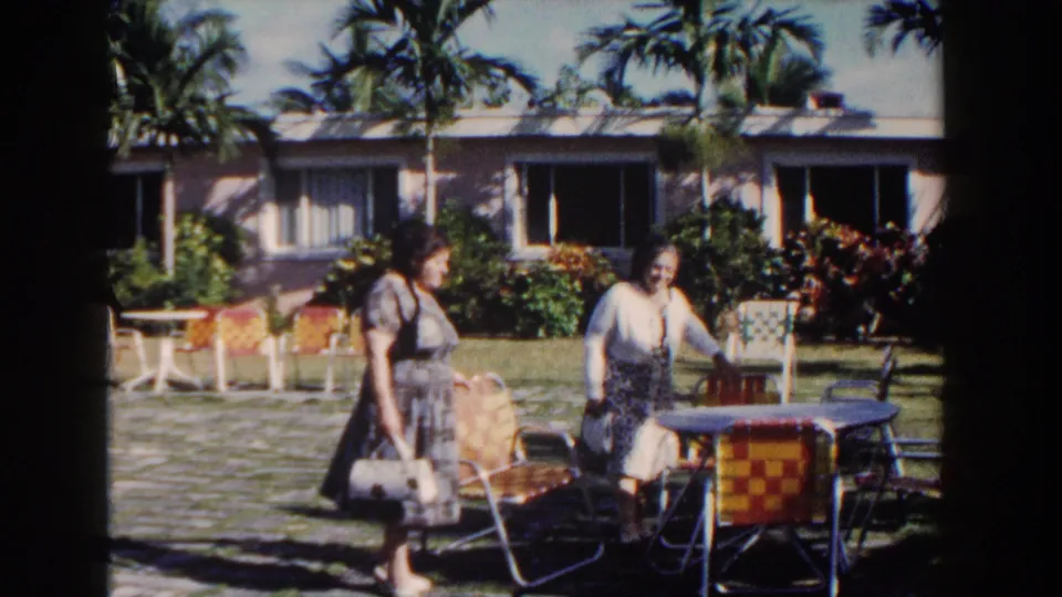In the Nassau resort courtyard, the women move past woven chairs and a small table, with tropical plants and a low pink building behind them.