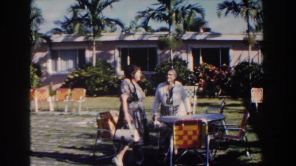 Near the end of the clip, the women appear to slow beside a small table and woven chairs in the 1961 Nassau courtyard, with palms and resort windows behind them.
