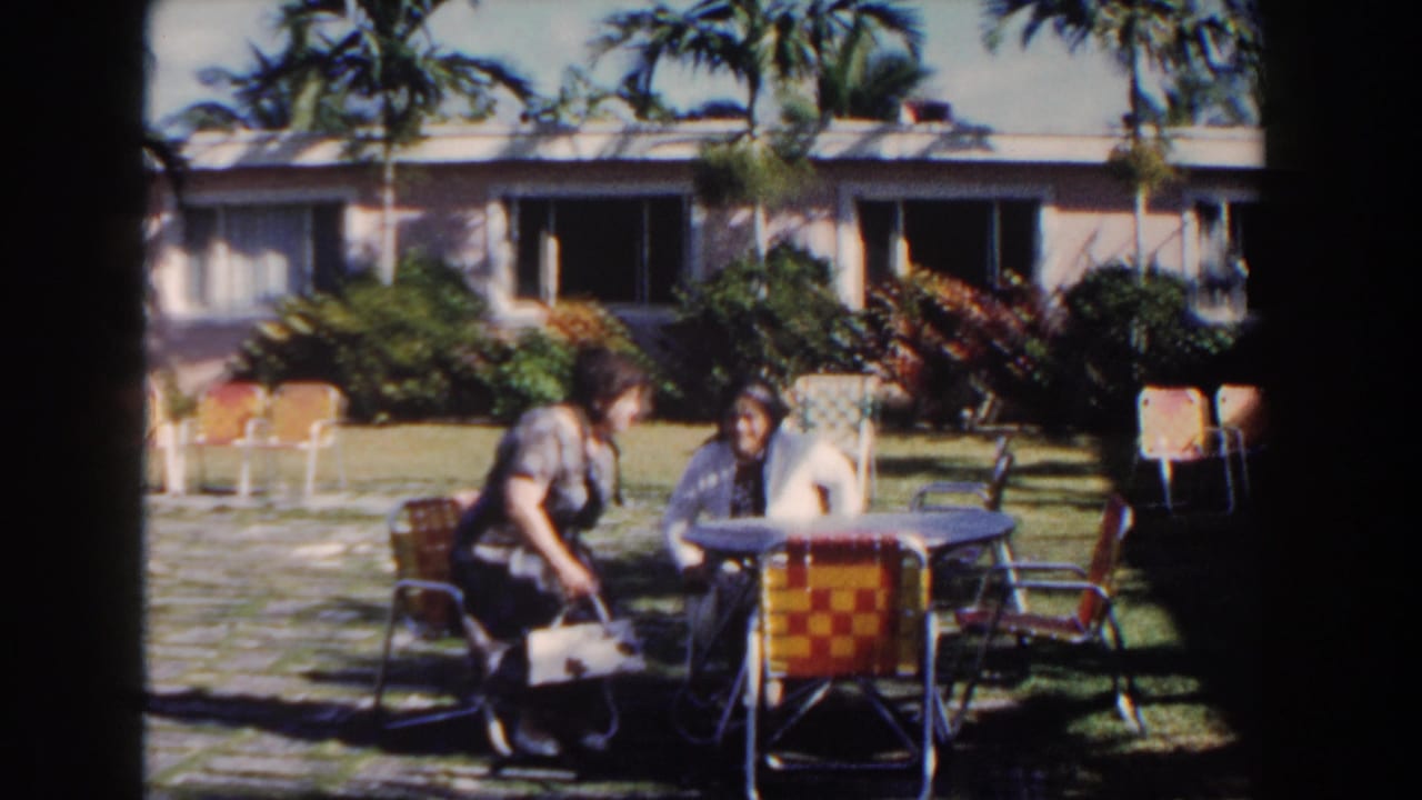 Two women sit by a patio table in a sunny tropical garden.