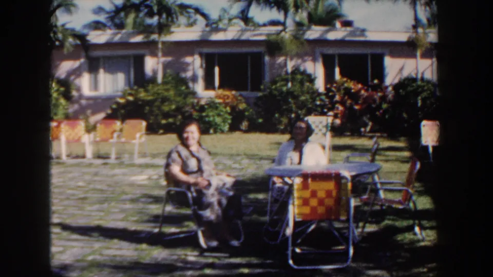 Two seated women appear relaxed at a garden table in 1961 Nassau, with colorful chairs, palms and a low house in the background.